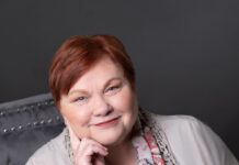 Woman sits in chair with one hand on side of her head and looks at the camera.