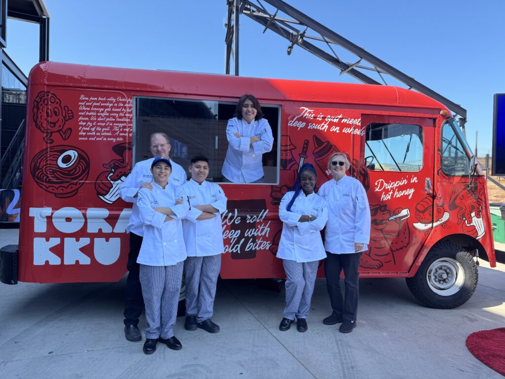 Several individuals wearing white chef uniforms stand in front of a red food truck, while one individual also wearing a white chef uniform stands at the window from inside the food truck.
