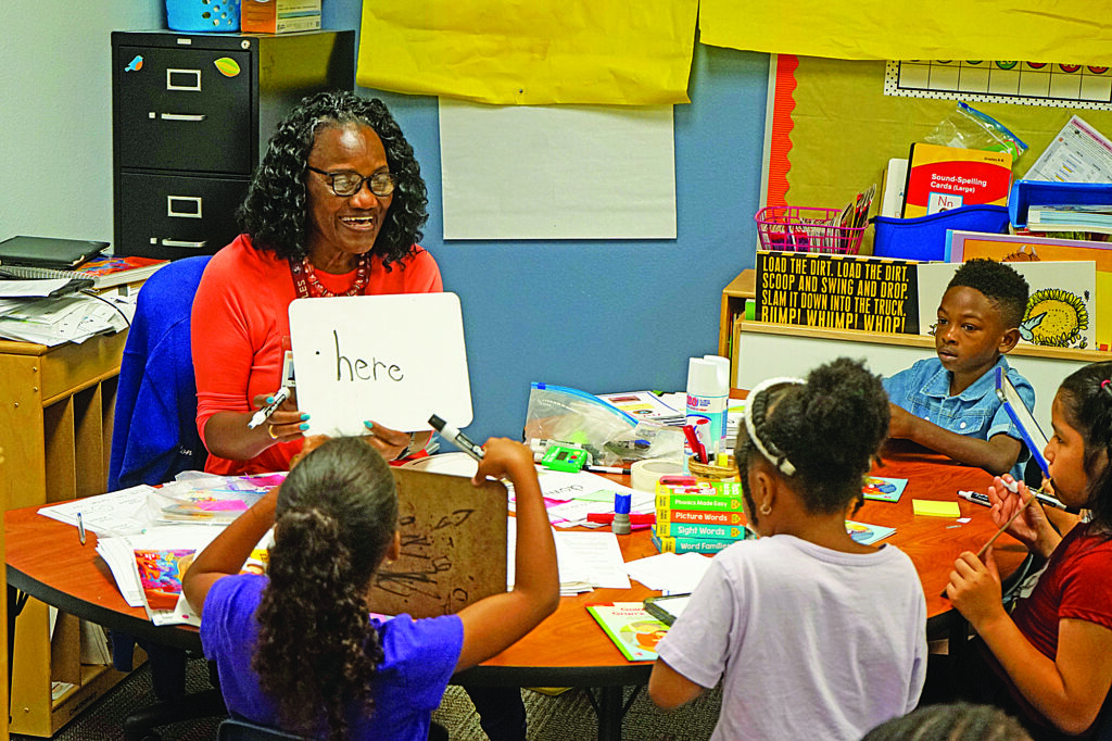 Kindergarten teacher Betty Washington holds a small whiteboard with the word "here" written on it to a table full of children during a lesson.