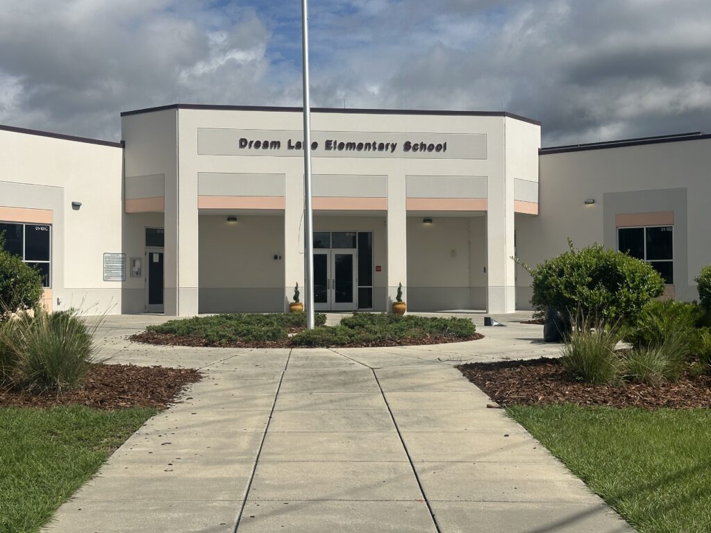 A gray-and-white building with the words "Dream Lake Elementary School" written across its front, with a flag pole in front of the building and a pathway leading up to the building.