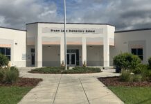 A gray-and-white building with the words "Dream Lake Elementary School" written across its front, with a flag pole in front of the building and a pathway leading up to the building.