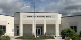 A gray-and-white building with the words "Dream Lake Elementary School" written across its front, with a flag pole in front of the building and a pathway leading up to the building.
