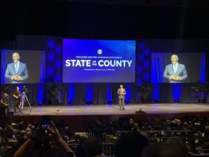 Man stands on stage with three large TV monitors behind him. The center monitor shows words "State of the County." The two monitors on either side of the center monitor show image of the man standing on stage.