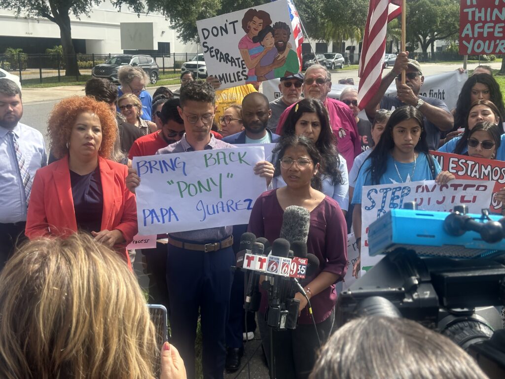 Several people in front of a microphone hold protest signs and wave an American flag.