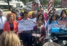 Several people in front of a microphone hold protest signs and wave an American flag.