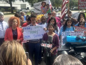 Several people in front of a microphone hold protest signs and wave an American flag.