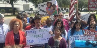 Several people in front of a microphone hold protest signs and wave an American flag.