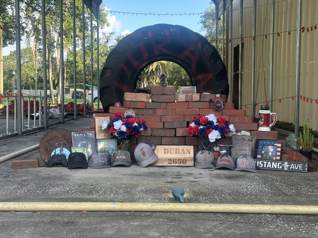 A memorial consisting of a large tire with the name "DURAN" painted on it behind a stack of bricks. IN front of the stack of bricks are caps, flowers, and video game cases. A wooden placard says "Duran 2650" is placed in the bottom center, in front of the brick stack.