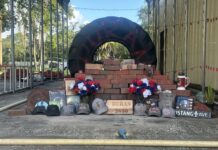 A memorial consisting of a large tire with the name "DURAN" painted on it behind a stack of bricks. IN front of the stack of bricks are caps, flowers, and video game cases. A wooden placard says "Duran 2650" is placed in the bottom center, in front of the brick stack.