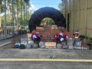 A memorial consisting of a large tire with the name "DURAN" painted on it behind a stack of bricks. IN front of the stack of bricks are caps, flowers, and video game cases. A wooden placard says "Duran 2650" is placed in the bottom center, in front of the brick stack.