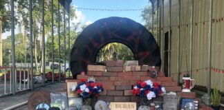 A memorial consisting of a large tire with the name "DURAN" painted on it behind a stack of bricks. IN front of the stack of bricks are caps, flowers, and video game cases. A wooden placard says "Duran 2650" is placed in the bottom center, in front of the brick stack.