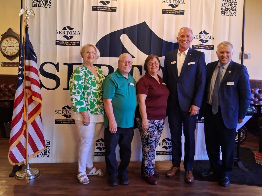 Five people -- two women and three men -- stand in front of the Sertoma Club seal, with the U.S. flag to the left.