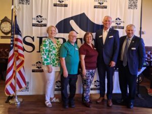 Five people -- two women and three men -- stand in front of the Sertoma Club seal, with the U.S. flag to the left.