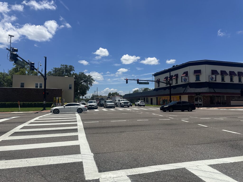 An intersection with white lines on the asphalt road, cars on the road, and buildings in the background.