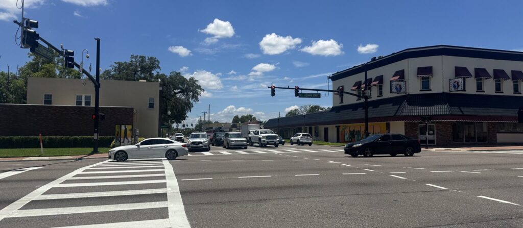 An intersection with white lines on the asphalt road, cars on the road, and buildings in the background.