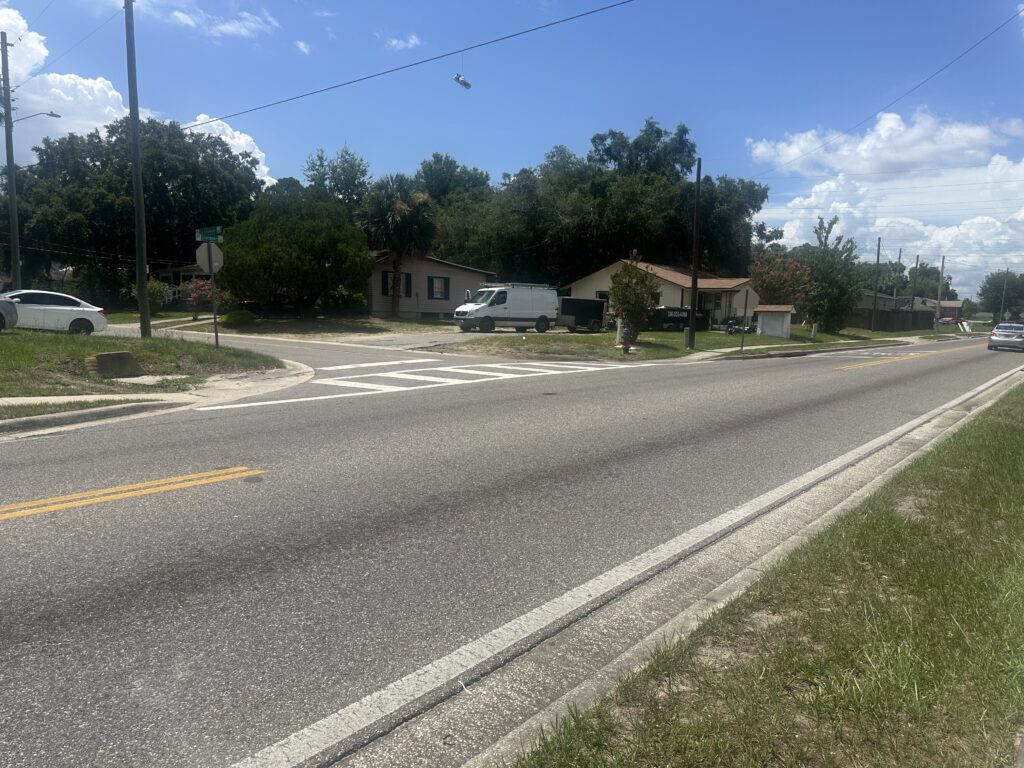 An asphalt road stretches across the bottom of the photo, with another asphalt road intersecting it. A house and vehicles are in the background.