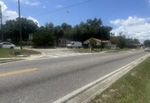 An asphalt road stretches across the bottom of the photo, with another asphalt road intersecting it. A house and vehicles are in the background.