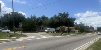 An asphalt road stretches across the bottom of the photo, with another asphalt road intersecting it. A house and vehicles are in the background.