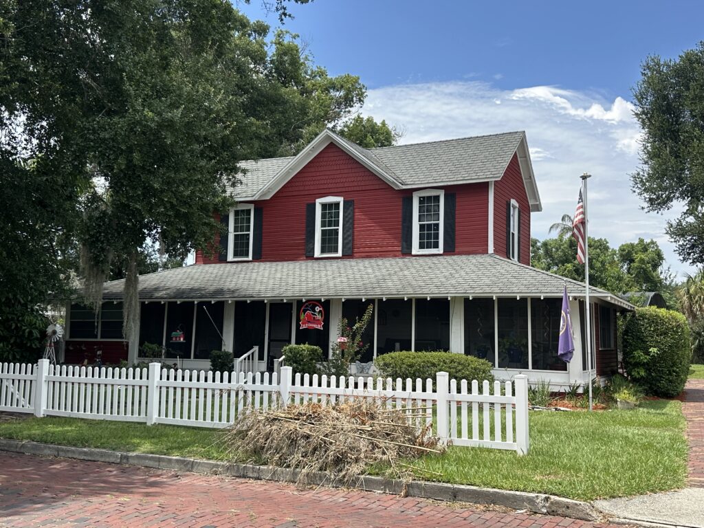 A two-story red house with gray roofs and black-and-white framed windows. A picket white fence and a brick road are in front of this house. Trees surround the house.