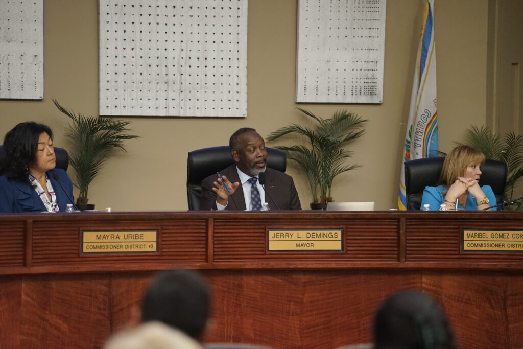 A man and two women sit on a dais, with a desk in front of them. A plaque with each individual's name is mounted on the desk.