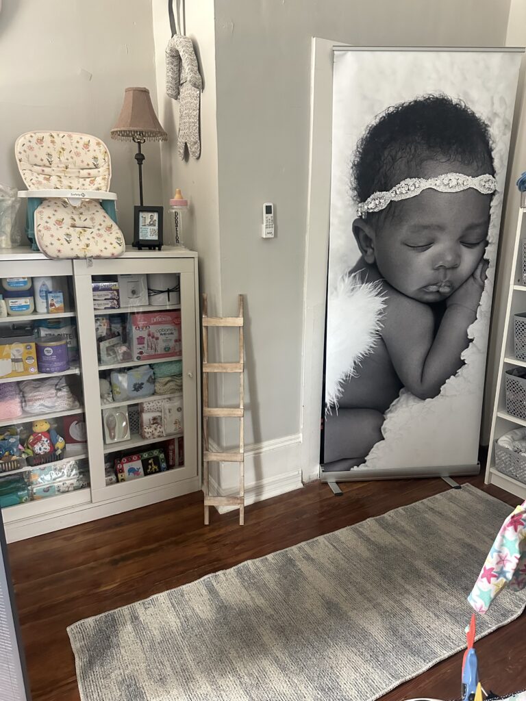 A room with a cabinet stocked with baby supplies and a large picture of a baby on the right side. A rug is on the floor.
