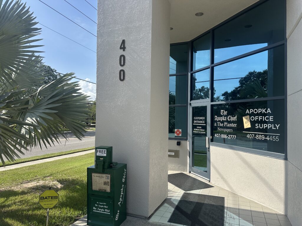 An office building with the number 400 going horizontally down a column. A newspaper vending box stands in front of this column. The words "The Apopka Chief and The Planter" and "Apopka Office Supply" appear on the front window of the office.