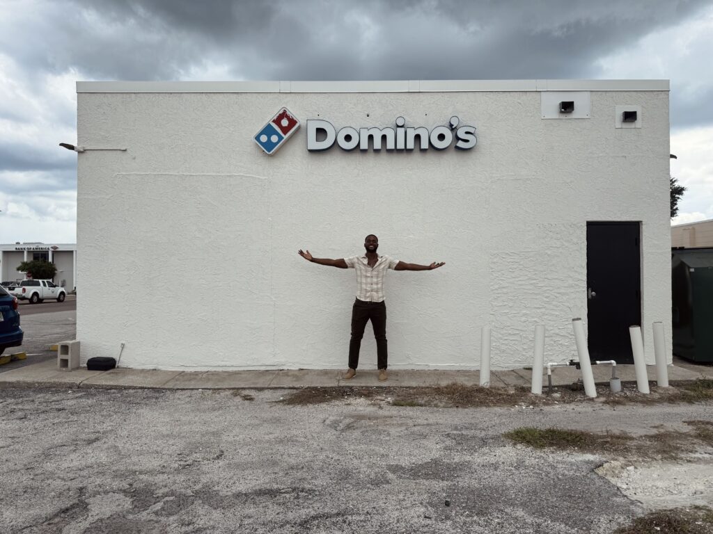 A man stands in front of a Domino's building. The building is painted white, with the word Domino's over the man's head.