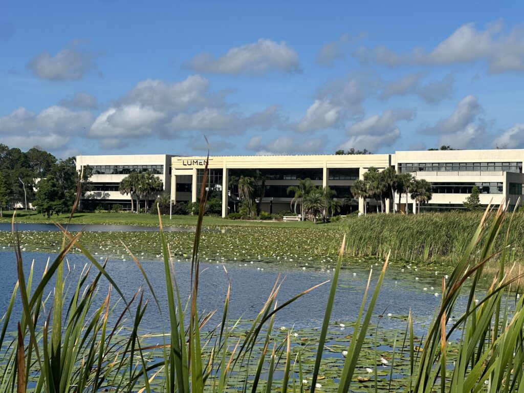 A blue lake surrounded by green grass with an office building in the background and blue sky with clouds looming overhead.