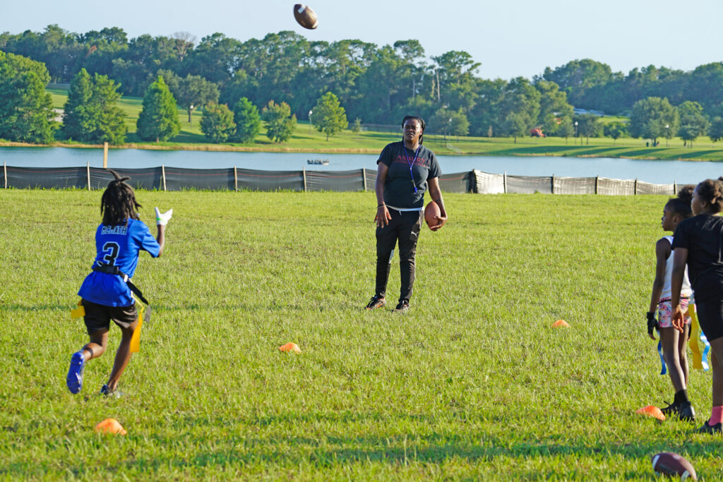 A young girl catches a pass from her coach at flag football practice in a field in front of a lake.