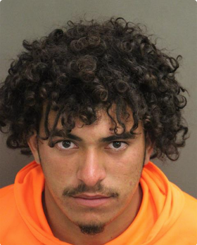Mug shot of young man with black curly hair, mustache and wearing an orange shirt.