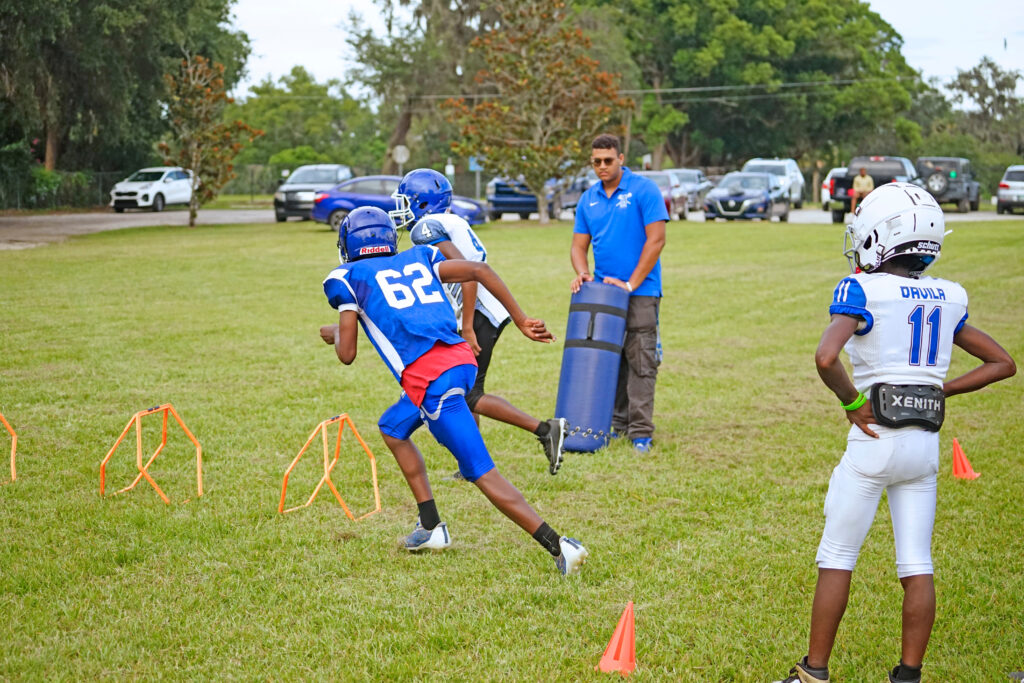 Two young kids in football gear run through a drill with an older player watching.