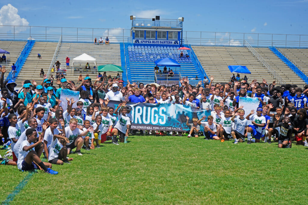 A group of kids and adults stands behind and next to a sign for drug awareness.