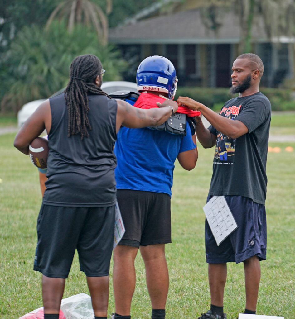 Two coaches help a Pop Warner player with his pads.