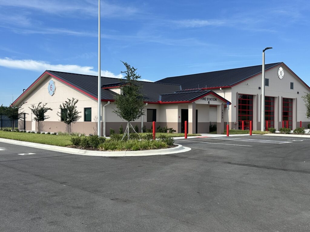 Apopka Fire Station 6, with pavement in front of it and blue sky behind it. The building itself has a dark gray roof and off-white exterior paint color.