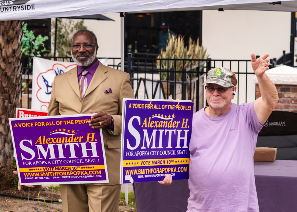 John Cloran (right) waves alongside Commissioner Alexander Smith (left) outside the Apopka Community Center.