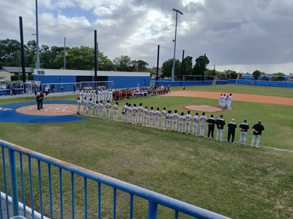 The Apopka Blue Darters and Olympia Titans stand for the National Anthem along with dozens of Little Leaguers along the third base line.