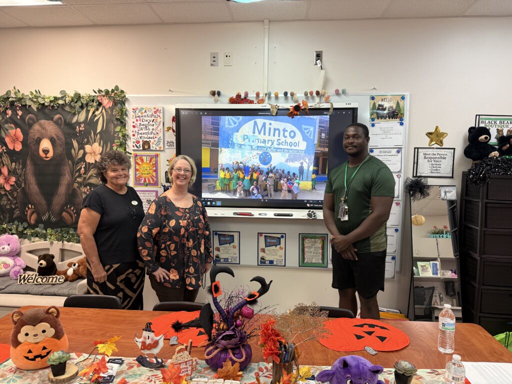 From left to right: GFWC Apopka Woman's Club President Diane Harrison, Clay Springs Elementary School Partners in Education Coordinator Christine Muchow, and Clay Springs P.E. coach Aden Jackson pose with a picture of Minto Primary School students receiving the equipment.